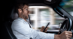 A man is driving his car with an expression of shock. His phone is seen in his hand.